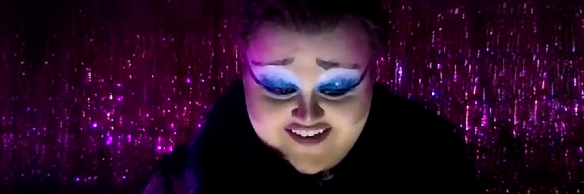a woman in heavy theatrical makeup in front of a sparkly pink background